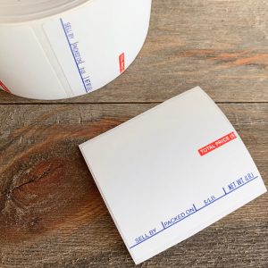 A roll of blank white vintage butcher price label stickers with a loose sticker next to it, showing the little bit of blue and red text